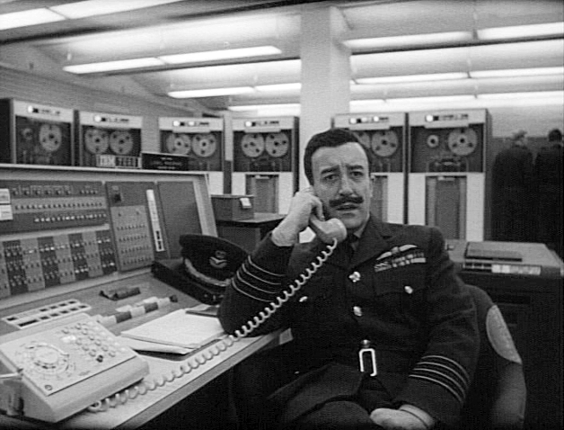 Peter Sellers as Group Captain Lionel Mandrake in Stanley Kubrick's 1964 film, Dr. Strangelove. Sellers is sitting at the console of an w:IBM 7090 computer with w:IBM 729 magnetic tape drives in the background.
