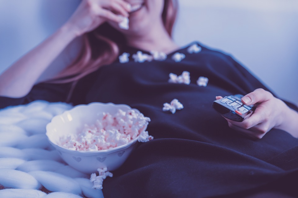 a girl holding a remote, white bowl with a popcorn, popcorn spilling on the girl’s shirt