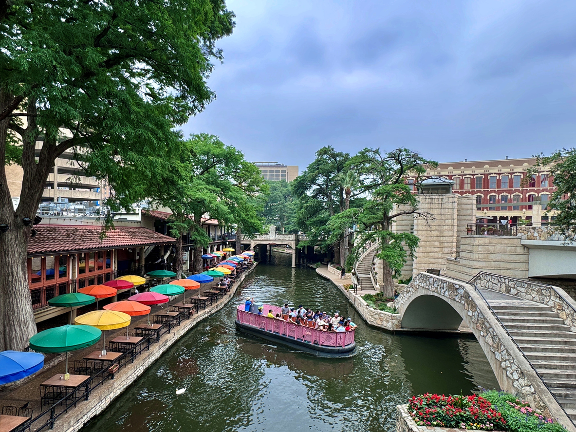 A scenic view of the San Antonio River Walk featuring a riverboat cruise passing under historic bridges